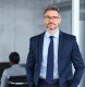 Portrait of handsome mid adult business man standing in modern office. Successful mature entrepreneur in formal clothing looking at camera with satisfaction. Confident man in suit with eyeglasses and beard standing with hands in pocket and looking at camera in office.